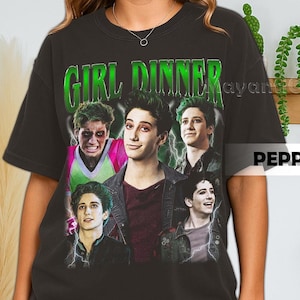 May include: Black t-shirt featuring a graphic with the text "GIRL DINNER" in green, with images of several young men. The design includes lightning bolts and a vintage aesthetic.