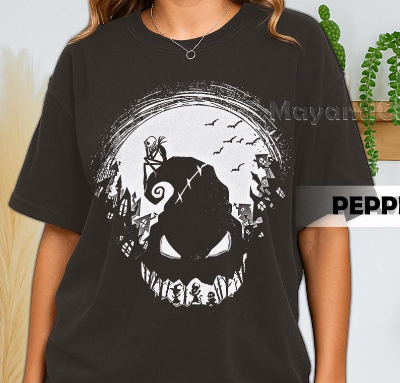 K&ouml;nnte beinhalten: Dunkelgraues T-Shirt mit einem wei&szlig;en Aufdruck eines gruseligen Charakters mit breitem Grinsen, vor einem Vollmond mit Flederm&auml;usen und einer Stadtsilhouette. Das Shirt hat die Texte "Mayan" und "PEPPI".