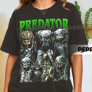 Könnte beinhalten: Schwarzes T-Shirt mit einem Grafikdesign von mehreren Predator-Charakteren. Das Wort "PREDATOR" ist in grünen Blockbuchstaben gedruckt. Das Design zeigt detaillierte Illustrationen der außerirdischen Jäger, die ihre Masken und Waffen präsentieren.