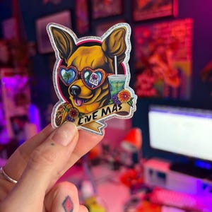 May include: A sticker featuring a chihuahua wearing heart-shaped sunglasses with palm trees and a building design. The dog holds a drink and a taco, with a banner that reads "LIVE MAS". The sticker has a glittery border.