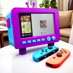 Könnte beinhalten: Eine lila und blaue Gaming-Konsole im Retro-Stil mit einem Bildschirm, der Spieloptionen anzeigt. Zwei Controller, einer blau und einer orange, befinden sich vor der Konsole. Die Konsole hat ein Fernsehdesign mit antennenartigen Merkmalen.