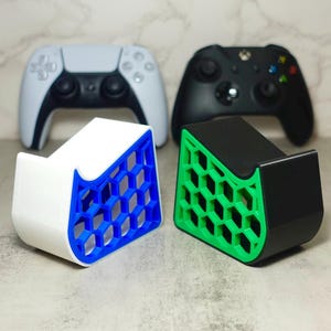 May include: Three 3D printed game controller stands in white, black, and green with a honeycomb pattern. The stands are designed to hold a video game controller upright.