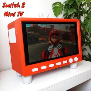 May include: An orange Mini TV with the text "Switch 2 Mini TV" on top. The screen shows a video game character in a red kart. The TV has white buttons and feet.