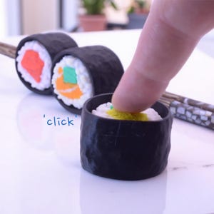 Puede incluir: Un conjunto de tres rollos de sushi negros con rellenos coloridos, que incluyen naranja, verde y amarillo. Un dedo presiona un cuarto rollo de sushi, que está en un tazón negro. El texto 'click' está escrito debajo del tazón.