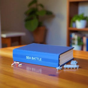 Può includere: Una scatola di gioco blu con la scritta "SEA BATTLE" è su un tavolo di legno. Navi giocattolo in miniatura e pedine sono accanto alla scatola. La scatola del gioco è rettangolare e ha pagine bianche sul lato.