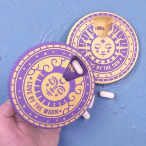May include: A purple and gold pill organiser with a sun and moon design. The organiser has the text "LOVE BY THE MOON" and "LIVE BY THE SUN". The organiser is holding pills and has the text "Mo".