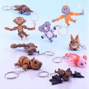 May include: A collection of animal-shaped keychains, including a platypus, monkey, orangutan, koala, rabbit, squirrel, capybara, axolotl, and bat. Each keychain is made of a light brown, gray, orange, and pink material and features a silver keyring.