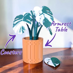 Pode incluir: Um vaso de plantas laranja decorativo com um design de folha de Monstera verde e branca. O vaso é cilíndrico com detalhes de ripas verticais. Uma coaster correspondente está ao lado do vaso. O texto na imagem diz "Coasters" e "Armrest Table".