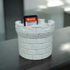 Puede incluir: Un soporte blanco en forma de castillo para un cartucho de juego de Nintendo Switch. El soporte está hecho de un material texturizado y tiene un patrón similar a un ladrillo. Un cartucho de juego de Nintendo Switch está insertado en el soporte.