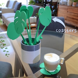 Può includere: Sottobicchieri a forma di foglia di monstera verde con una tazza di caffè su uno dei sottobicchieri. I sottobicchieri sono in un vaso con altri sottobicchieri a forma di foglia di monstera.