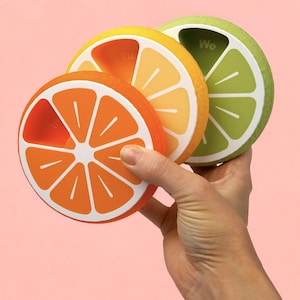 May include: Three colourful, citrus-themed pill organisers. The orange, yellow, and green organisers are shaped like orange and lime slices, with white details. Each has a section labelled with a day of the week. A hand is holding the organisers.