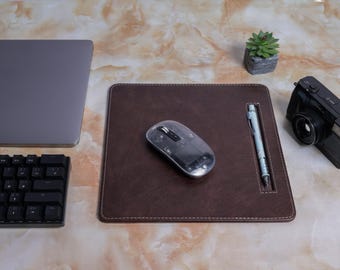 May include: A workspace setup featuring a brown leather mousepad with a wireless mouse and a pen. A laptop, keyboard, and camera are also present, along with a small potted succulent. The items are arranged on a marble-patterned surface.