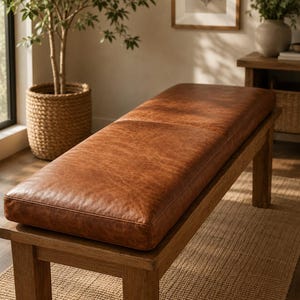 May include: A rectangular bench featuring a smooth, cognac-colored leather seat cushion with visible grain texture, supported by a sturdy wooden frame with four straight legs. The seat is long and flat, providing a clean, horizontal surface.
