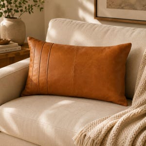 May include: A rectangular lumbar pillow in a solid cognac brown color. The pillow features a smooth, leather-like texture with three vertical stitched seams positioned on the left side, creating a clean, structured appearance on the cushion.