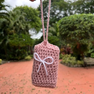 May include: A small, handmade, pink crochet pouch with a white bow detail. The pouch is suspended by a matching pink cord. The background features lush green foliage and a blurred outdoor setting.