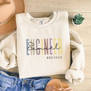 May include: A cream-coloured sweatshirt with the word "Engineer" in colourful vertical stripes and the name "Samuel" written in cursive below. The text "EST. 2024" is printed below the name.