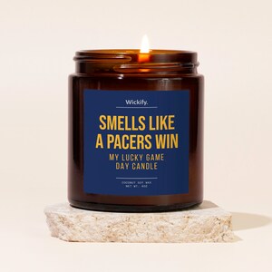 May include: A brown glass candle jar with a blue label that reads "Wickify. Smells Like A Pacers Win My Lucky Game Day Candle Coconut Soy Max Net Wt. 4 oz". The candle is lit and the flame is visible.
