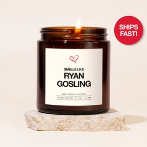 May include: A brown glass candle jar with a white label that reads "Smells Like Ryan Gosling" with a red heart symbol above the text. The candle is lit and the label also includes the text "Hand Poured by Wickify. Coconut Soy Wax 4 oz / 20 hrs".