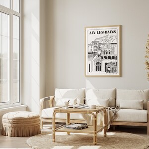 May include: A living room interior with a white couch, a wooden coffee table, a wicker ottoman, and a framed black and white print of a city skyline with the text "Aix-les-Bains".