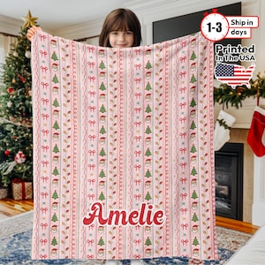 May include: A pink Christmas-themed blanket with a repeating pattern of Santa Claus faces, Christmas trees, bows, and stars. The name "Amelie" is printed in red script. The blanket is printed in the USA.