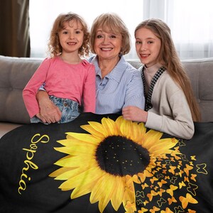 May include: A black fleece blanket featuring a large yellow sunflower and a scattering of yellow butterflies. The blanket is personalised with the name "Sophia" written in yellow script on the left side.