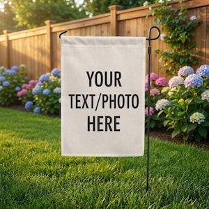 May include: A rectangular off-white garden flag hanging from a black metal stand. The flag features the black text YOUR TEXT/PHOTO HERE centered on the front. The fabric has a visible weave texture and is displayed outdoors in a green yard.