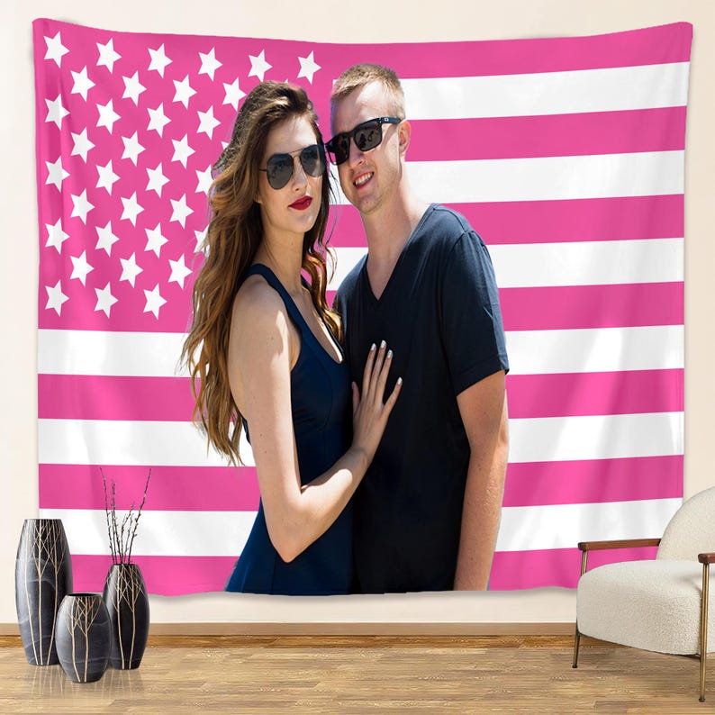 May include: A pink and white American flag tapestry with a couple posing in front of it. The woman wears a dark blue dress and sunglasses, and the man wears a black shirt and sunglasses. The tapestry has white stars and pink stripes.
