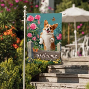 May include: A garden flag featuring a smiling corgi dog with a welcoming sign. The flag has a teal background with pink roses and butterflies. The sign says "welcome".