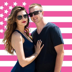 May include: A couple wearing sunglasses poses in front of a pink American flag background. The woman wears a navy blue dress and the man wears a navy blue v-neck shirt. The woman has long wavy brown hair and red lipstick.