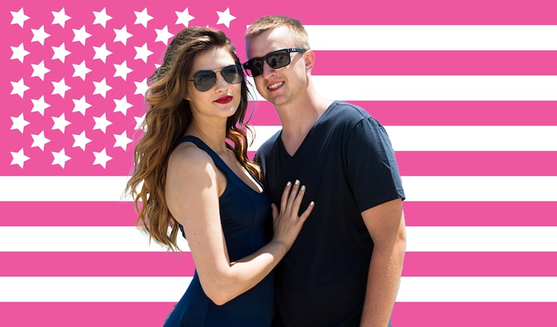 May include: A couple wearing sunglasses poses in front of a pink American flag background. The woman wears a navy blue dress and the man wears a navy blue v-neck shirt. The woman has long wavy brown hair and red lipstick.