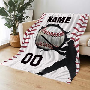 May include: White baseball-themed blanket featuring a baseball bursting through a wall, a baseball bat, and a silhouette of a baseball player. The blanket has the word "NAME" and the number "00" printed on it.