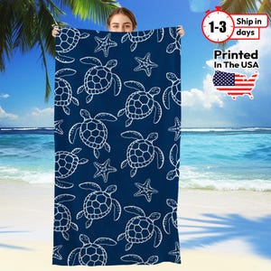 May include: A navy blue beach towel featuring a white turtle and starfish pattern. The towel is held up against a beach backdrop with palm trees and ocean waves. The text "Printed In The USA" is visible.
