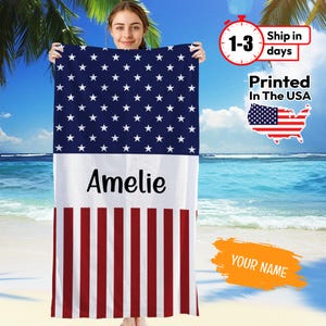 May include: A personalized beach towel featuring the American flag design. The top portion has a blue background with white stars, the middle section is white with the name "Amelie", and the bottom has red and white stripes. The towel is printed in the USA.