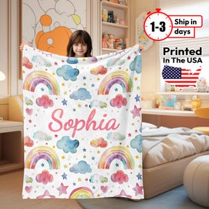 May include: White blanket with a watercolor design of rainbows, clouds, stars, and hearts. The name "Sophia" is printed in pink script. The blanket is printed in the USA.