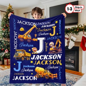 May include: A navy blue personalized blanket featuring the name "Jackson" in white and yellow, along with illustrations of construction vehicles. The design includes a crane, dump truck, excavator, and cement mixer. The blanket has the text "1-3 Ship in days".