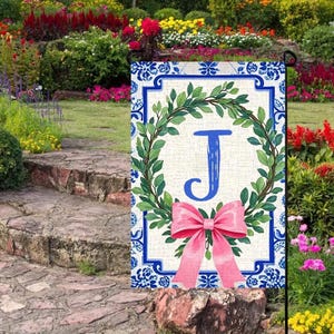 May include: A garden flag featuring a blue letter "J" inside a green leaf wreath, with a pink bow at the bottom. The flag has a blue and white floral border. The flag is displayed in a garden setting with colorful flowers.