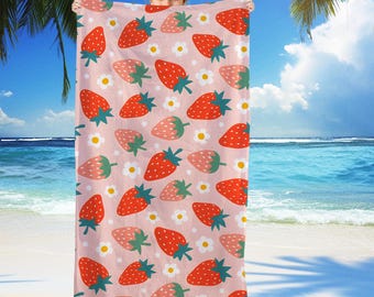 May include: A pink beach towel with a repeating pattern of red and pink strawberries and white daisies. The towel is held up against a beach backdrop with palm trees and blue water.