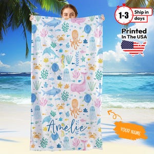 May include: White beach towel featuring a colorful sea life pattern with whales, octopuses, and jellyfish. The towel has the name "Amelie" in cursive script. Printed in the USA.