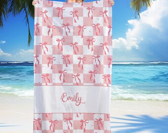 May include: A pink and white checkered beach towel with pink bow accents. The towel has a white band with the name "Emily" in cursive script. The towel is held up against a beach background.
