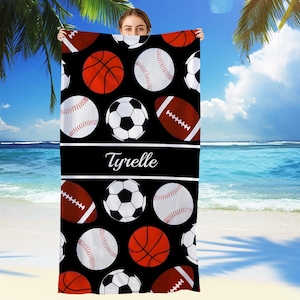 May include: Black beach towel featuring a pattern of red basketballs, baseballs, soccer balls, and footballs. The towel has a black stripe with the name "Tyrelle" in white script. The background is a beach scene with palm trees and blue water.