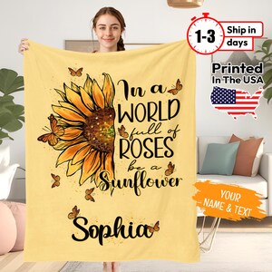 May include: A yellow fleece blanket featuring a sunflower graphic and the text "In a world full of roses be a sunflower". The name "Sophia" is printed below the graphic.
