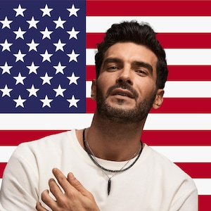 May include: A person wearing a white crew neck shirt and a black beaded necklace with a black pendant. The background features the American flag with red and white stripes and a blue field with white stars.