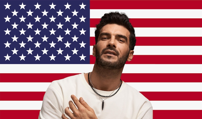 May include: A person wearing a white crew neck shirt and a black beaded necklace with a black pendant. The background features the American flag with red and white stripes and a blue field with white stars.