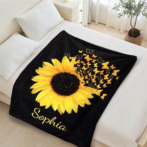 May include: Black fleece blanket featuring a yellow sunflower with a scattering of yellow butterflies. The name "Sophia" is printed in yellow script below the sunflower.