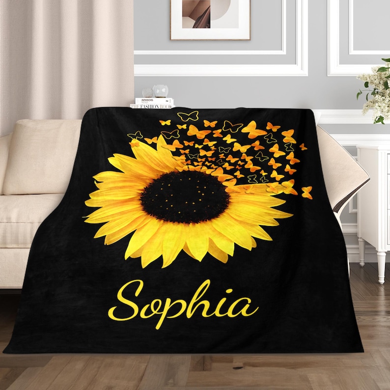 May include: Black fleece throw blanket featuring a yellow sunflower with butterflies flying away from it. The name "Sophia" is written in yellow cursive below the sunflower.