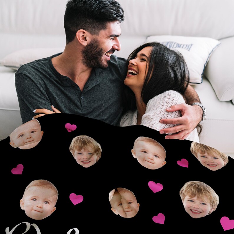 May include: A black fleece blanket with a repeating pattern of pink hearts and faces of children. The blanket is being held by a couple on a couch.