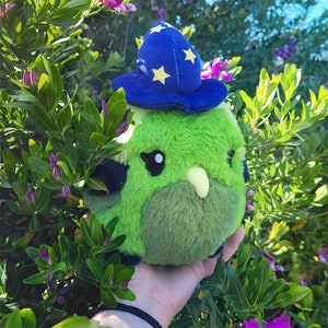 May include: A plush, round, green creature wearing a blue wizard hat with yellow stars. The creature has black eyes, a light green beak, and a darker green belly. The plush is held in front of a background of green leaves and purple flowers.