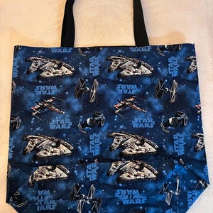 Può includere: Borsa tote blu con un design di Star Wars con astronavi e la scritta bianca "STAR WARS". La borsa ha manici neri ed è realizzata in tessuto resistente.