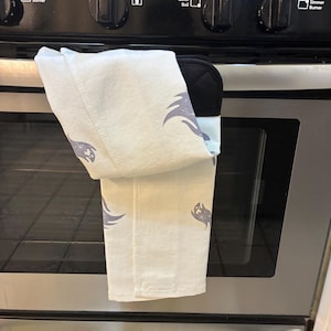 Puede incluir: Un pa&ntilde;o de cocina blanco y un guante de horno negro cuelgan de un horno de acero inoxidable. La toalla tiene un patr&oacute;n repetido de un dise&ntilde;o gris estilizado. El horno tiene perillas de control negras y una puerta de vidrio.