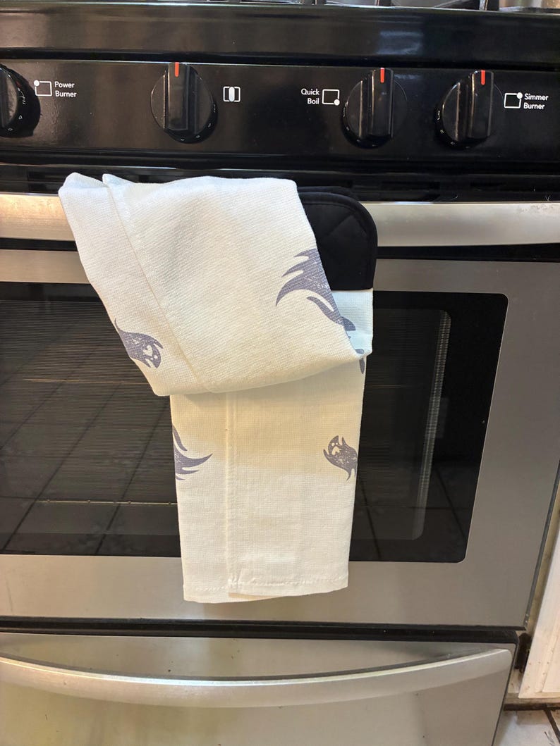 Puede incluir: Un pa&ntilde;o de cocina blanco y un guante de horno negro cuelgan de un horno de acero inoxidable. La toalla tiene un patr&oacute;n repetido de un dise&ntilde;o gris estilizado. El horno tiene perillas de control negras y una puerta de vidrio.